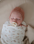 Newborn baby swaddled in a Pink Flowers swaddle blanket with a Cedar bonnet and Cream Frigg Daisy rubber pacifier, lying on a soft surface.