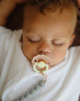 Lifestyle image of a baby sleeping with a Frigg Daisy Night pacifier in a white shirt