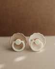 Two Frigg Daisy Night pacifiers on a beige surface
