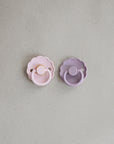 Two Frigg Daisy Rubber pacifiers, one Baby Pink and one Soft Lilac, on a light gray background