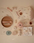 A flat lay featuring pastel baby essentials, including a Mushie Classic Silicone Suction Plate, bib, sippy cup, pacifier, teether, snack container, cup, and a folded floral-patterned blanket tied with ribbon.