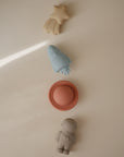 Four pastel silicone bath toys from mushie's Space Mold Free Bath Play Set—a yellow shooting star, blue rocket, red disc, and light gray astronaut—are arranged vertically on a beige background with diagonal light.