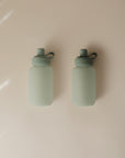 Two identical mushie Silicone Puree Pouches in matte light green, each with a screw top and loop handle, are placed side by side on a smooth light beige background, highlighting their mess-free feeding design.