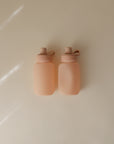 Two small, square mushie Silicone Puree Pouches in peach, each with a round cap and looped handle, sit side by side on a light beige surface. Soft natural light highlights the reusable design for convenient mess-free feeding.