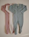 Three mushie Zipper Pajamas with feet in dusty rose, cream, and light teal are laid out side by side on a neutral background. These cozy long-sleeve onesies offer soft, comfortable sleepwear for babies.