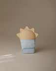 The mushie Sun Teething Mitten is a light blue silicone glove with a beige, sun-shaped baby teether on top, shown standing upright against a neutral beige background.