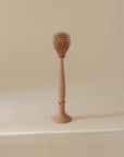 The mushie Silicone Baby Bottle Brush, featuring spiky silicone bristles and an oval head, stands upright in its matching holder against a plain beige background.