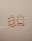 Two light pink, arch-shaped No-Drop Sensory Teethers by mushie, featuring textured knobs and bars, are displayed on a beige surface—ideal as a sensory teether for your baby against a plain background.