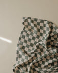 The mushie Organic Cotton Muslin Swaddle Blanket, featuring a green and white checkered pattern, is displayed crumpled on a plain beige surface, softly lit with a faint streak of light on the left side.