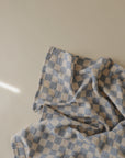 The mushie Organic Cotton Muslin Swaddle Blanket, featuring a soft blue and white checkered pattern, is displayed on a light beige surface and bathed in gentle natural light from the left.