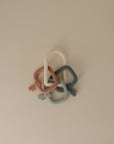 The mushie Key Toy features four interlocking silicone teething rings in pastel pink, white, green, and blue, arranged on a beige surface—perfect for gently soothing babies’ gums.