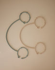 The mushie Adjustable Silicone Strap 2-Pack features blue and beige spiral cords, each forming a semicircle between matching round rings in a mirrored pattern on a beige background—ideal as baby essentials.