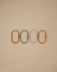 The mushie Chain Link Rings include four oval-shaped plastic links in red, green, white, and yellow, neatly arranged on a beige background—ideal for promoting fine motor skills during play.