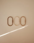 Three oval-shaped mushie Chain Link Rings in muted beige and brown tones are arranged side by side on a light surface, with a diagonal beam of light crossing the lower left corner.