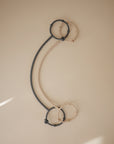 Two curved, spiral adjustable silicone straps—one dark gray and one beige—from mushie’s Adjustable Silicone Strap 2-Pack are linked by four circular rings at each end and set against a neutral beige background with soft light from the lower left.