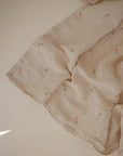 The mushie Organic Cotton Muslin Swaddle Blanket, in light beige with small colorful embroidered motifs, features a delicate, slightly wrinkled texture and gentle folds, softly displayed on a smooth cream-colored surface.