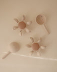 Four cream-colored mushie Water Wheel Bath Toys with brown centers and petal-like, speckled handles are arranged on a light beige surface.