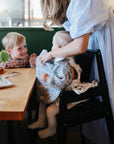 A woman in a light blue dress helps a baby in a patterned bib sit in a high chair at a wooden table. On the table, the mushie Water Resistant Wet Bag rests nearby, while a young child smiles at the baby amid food and a small vase.