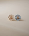 Two FRIGG Little Viking Silicone pacifiers by FRIGG, one cream and one soft blue, with round shields and tan silicone nipples, displayed side by side on a beige background. Made in Denmark.