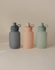 Three matte-finish water bottles in gray, peach, and light green with looped caps stand side by side on a beige background—ideal companions to your mushie Silicone Puree Pouch for mess-free feeding wherever you go.