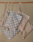 Four mushie Water Resistant Wet Bags hang from a wooden rod. The set features blue checkered, floral, and pale gold star patterns—ideal for diaper bag storage with zippered closures.