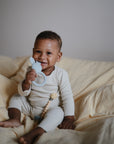 A smiling baby in the mushie Two Piece Set made from organic cotton sits on cream-colored sheets, holding a blue teething toy and a beaded pacifier clip.
