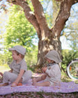 Two small children sit on a purple blanket under a tree in a sunny park. Both wear hats and light clothes, and one holds the mushie No-Drop Sensory Teether. A bicycle is parked nearby among pink flower petals on the ground.