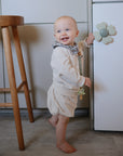 A smiling baby in a light outfit stands barefoot on the kitchen floor, holding a cabinet door with a flower knob. Beside the baby and stool are mushie Suction Spinner Toys, made from food-grade silicone for playful sensory fun.