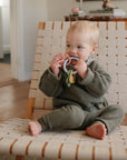 A toddler in a green knit outfit sits barefoot on a woven chair, chewing on the mushie Western Teething Ring in a cozy, sunlit room with wooden floors.