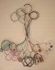 A set of pastel baby essentials from mushie, featuring the Adjustable Silicone Strap 2-Pack, rings, flower rattle, teether, palette toy, and animal shapes arranged symmetrically on a light background.