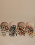 Four small beige containers tipped over, each spilling out mushie Chain Link Rings in muted green, blue, brown, and pink—displayed on a neutral background—perfect for activities that support fine motor skills.