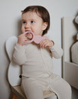 A toddler with short brown hair sits on a chair in a softly lit room, wearing mushie Zipper Pajama and holding a pink teething toy.