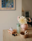 A wooden table displays a bowl with utensils, blackberries, a green water bottle, an ice cube tray, a mushie Silicone Puree Pouch for mess-free feeding, and a pink vase with flowers. Framed artwork is visible on the wall behind.