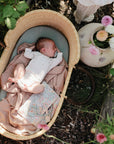 A baby sleeps in a basket lined with a pink blanket, surrounded by plants and flowers. On a small table, the mushie Water Resistant Wet Bag sits beside a vase of blossoms as sunlight filters through the leaves.