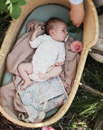 A baby in a white onesie lies in a wicker basket with a pink blanket, holding a floral cloth. Nearby, an adult hand holds a pink flower and the Mushie Diaper Cream Applicator amid greenery and blooming flowers.