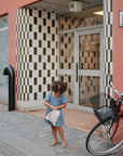 A young girl in a blue dress stands on a cobblestone sidewalk, holding a mushie Water Resistant Wet Bag in front of a building with a checkered tile entrance. A red bicycle with a basket is parked nearby.