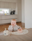 A baby smiles at a dining table in a modern kitchen, holding food while wearing the mushie Long Sleeve Bib. A bowl and sippy cup are on the table, with kitchen cabinets and appliances in the background.