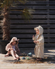 Two young children in animal-themed hooded bathrobes play on a sunny wooden deck. One stands and smiles, while the other sits near the mushie Dino Mold Free Bath Play Set, with a potted palm tree in the background.