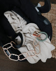 A black car seat holds a cream ribbed blanket and mushie Chain Link Rings, great for fine motor skill development, resting on a wooden floor next to a woven rug.