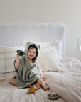 A young child sits on a bed wrapped in a hooded towel, smiling while holding mushie Bath Boats. Other stackable water play toys from mushie are scattered across the light-colored bedding.