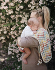 A blonde woman holds a baby outdoors before blooming pink flowers. The baby rests on her shoulder, with a mushie Organic Cotton Muslin Burp Cloth and pacifier clipped to it.