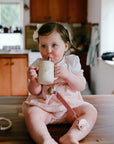 Toddler in pink uses Mushie Silicone Training Cup + Straw on the kitchen counter.