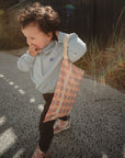 A young child with curly hair smiles outdoors on a sunny day, holding a mushie Water Resistant Wet Bag over one shoulder. Grass and a building are visible in the background.