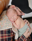 A sleeping newborn rests on a person in a plaid shirt and black cap, wrapped in a floral blanket, green knit wrap, and cuddled with mushie Organic Cotton Muslin Burp Cloth 2-Pack.