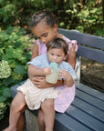 A young girl in a pink dress sits on a gray bench outdoors, holding a baby dressed in stripes. The baby enjoys mess-free feeding from the mushie Silicone Puree Pouch. Green plants and flowers decorate the background.