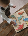 A young child stands at a wooden table, playing with the Mushie Wooden Puzzle, placing colorful dinosaur-shaped pieces onto the board to build fine motor skills.