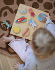 A young child sits on a patterned rug, playing with the Mushie Wooden Puzzle—an educational toy with space-themed shapes like an astronaut, rocket, planet, and moon—to help develop fine motor skills.