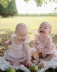 Two babies in pink dresses sit on a blanket outdoors, surrounded by apples and the mushie Princess Mold Free Bath Play Set. One baby smiles up in the sunlight while the other examines the play set toys.
