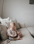 A baby in a light outfit plays with mushie Chain Link Rings on a beige sofa, surrounded by neutral pillows in a softly lit minimalist room—exploring toys that encourage fine motor skill development.