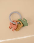 The mushie Western Teething Ring is made from food grade silicone, featuring a gray spiral handle and three animal-shaped charms in pink, yellow, and green, shown on a light beige background.